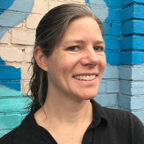 a white woman with brown hair and a black button-down shirt smiles in front of a mural in Little Five Points